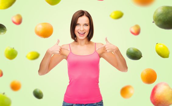 people, advertisement, diet, food and healthy eating concept - smiling woman in blank pink tank top pointing fingers to herself over fruits on green background