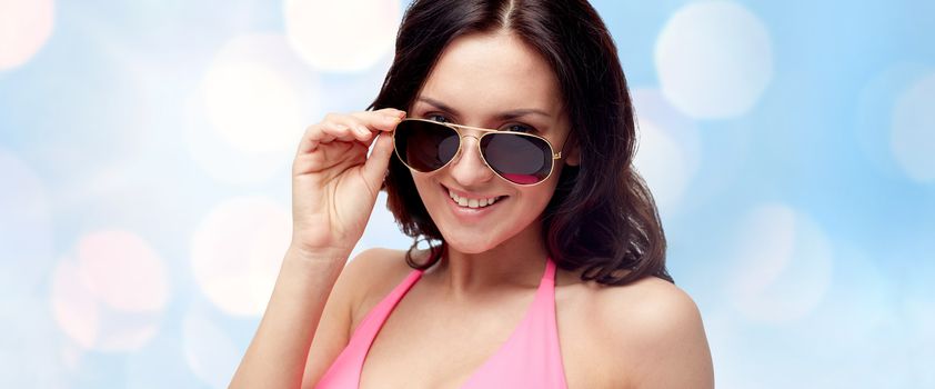 people, fashion, swimwear, summer and beach concept - happy young woman in sunglasses and pink swimsuit looking at you over blue holidays lights background