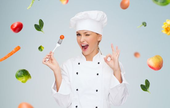cooking and food concept - smiling female chef, cook or baker with fork and tomato showing ok sign over falling vegetables on gray background