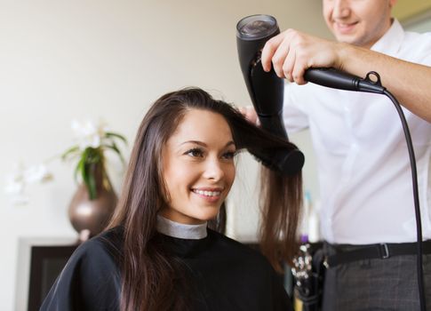 beauty, hairstyle and people concept - happy young woman and hairdresser with fan making hot styling at hair salon
