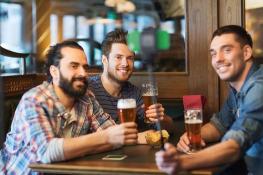 people, men, leisure, friendship and technology concept - happy male friends drinking beer and taking picture with smartphone selfie stick at bar or pub