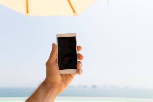 technology, travel, tourism, communication and people concept - close up of male hand holding smartphone with blank screen on summer beach