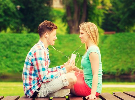 summer, vacation, holidays, technology and friendship concept - smiling couple with smartphone and earphones sitting on bench in park