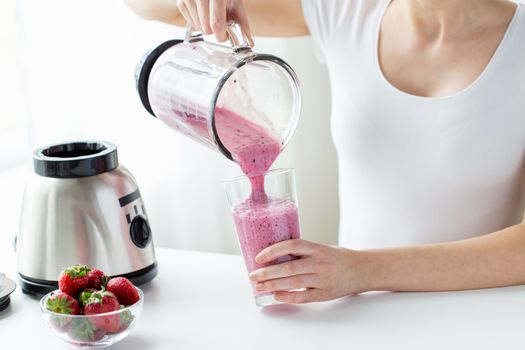 healthy eating, cooking, vegetarian food, dieting and people concept - close up of woman with blender and strawberries pouring milk shake to glass at home