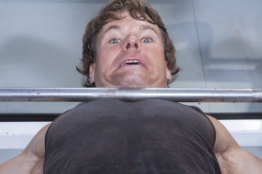 Closeup of helpless expression of Caucasian man stuck under heavy barbell on bench press at gym