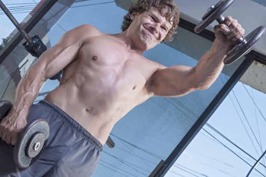 Muscular shirtless Caucasian man with expression of exertion as he raises heavy dumbbell weight while performing front dumbbell raises in gym