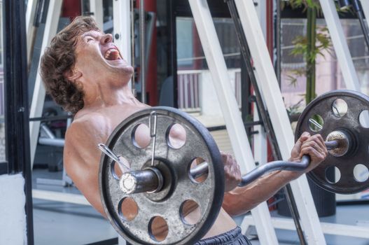 Shirtless muscular Caucasian man swings back and shouts while curling heavy weights on curl bar