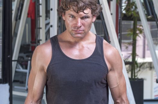 Muscular sweaty Caucasian man with intense expression looking at camera and wearing tank top in gym