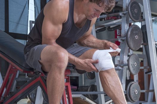 Muscular Caucasian man wraps knee with sport bandage in weightlifting gym