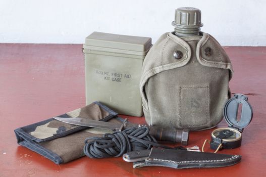 Assortment of survival hiking gear including first aid kit, canteen, knife, parachute cord, compass and map holder on old wood table