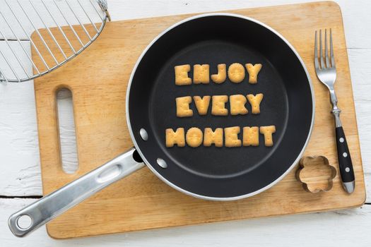 Top view of alphabet collage made of crackers. Quote ENJOY EVERY MOMENT putting in black pan. Other kitchenware: fork, cookie cutter and chopping board putting on white wooden table, vintage style image.