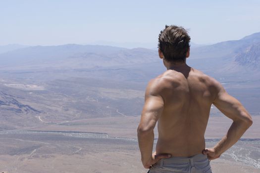 Muscular shirtless Caucasian man stands high on mountain top overlooking desert valley in Nevada