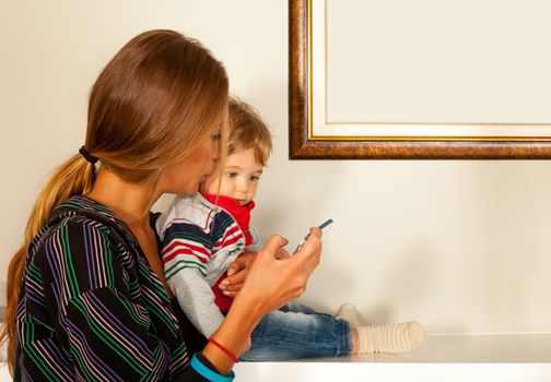 Young beautiful mother and a cute blue eyes child are looking at a smartfone indoors. Copy space frame.