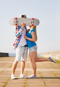 holidays, vacation, love and people concept - couple kissing and hiding their faces behind skateboard outdoors