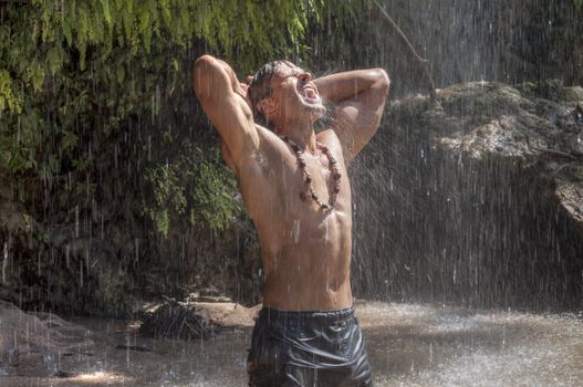 Handsome muscular shirtless Caucasian man stretches arms behind head and opens mouth to catch raindrops from waterfall in tropical paradise in Chiapas, Mexico