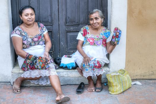 VALLADOLID, MEXICO - JANUARY 20, 2015: Two women in traditional dress sit in front of old rustic building along sidewalk offering textile handicrafts for sale to tourists
