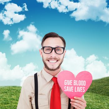 Geeky hipster smiling and holding heart card against blue sky over green field