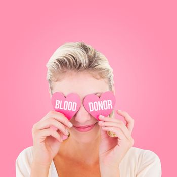 Attractive young blonde holding hearts over eyes against pink