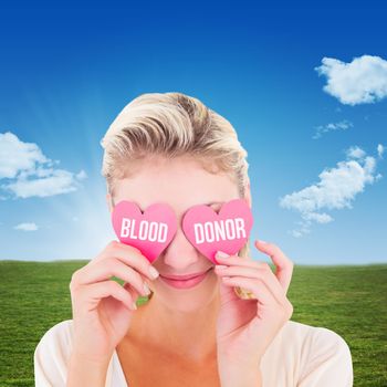 Attractive young blonde holding hearts over eyes against blue sky over green field