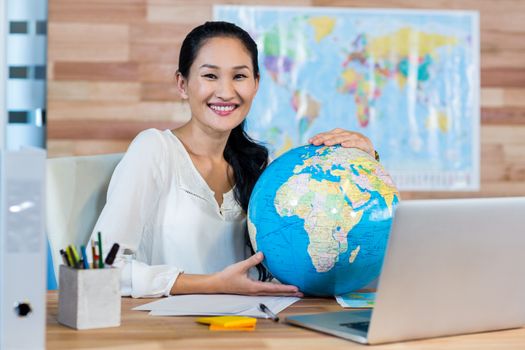 Pretty travel agent holding globe and smiling at camera in the office