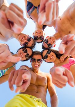 friendship, summer vacation, gesture and people concept - group of smiling friends wearing swimwear and pointing finger on you standing in circle over blue sky