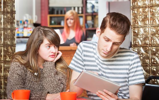 Sad woman looking over at boyfriend using computer tablet