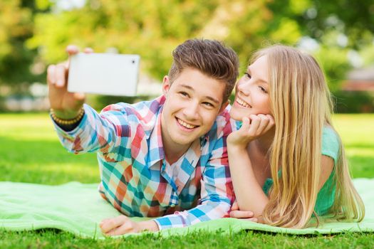 vacation, holidays, technology and friendship concept - smiling couple lying on blanket and making selfie with smartphone in park