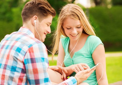 summer, vacation, holidays, technology and friendship concept - smiling couple with smartphone and earphones sitting in park