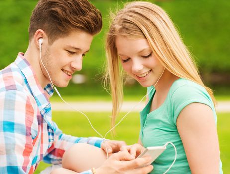 summer, vacation, holidays, technology and friendship concept - smiling couple with smartphone and earphones sitting in park