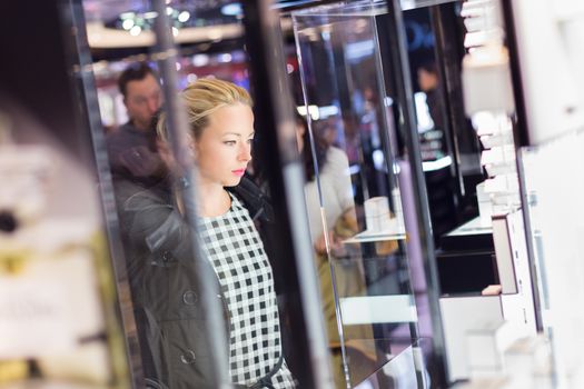 Beautiful blonde lady standing in front of showcase in beauty store, admiring new perfume collection.