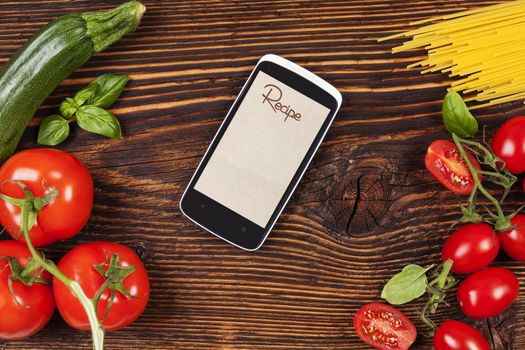 Fresh vegetables and pasta on wooden table with smartphone and recipe on screen. Groceries, recipe and cooking. Modern food preparation.