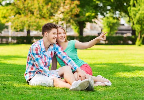 holidays, vacation, love and friendship concept - smiling couple sitting on grass and pointing finger in park