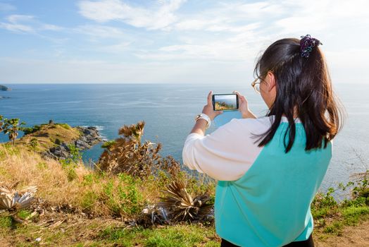 Female tourist taking photograph the island and sea on a high hill with smart phone at Cape Phromthep viewpoint a famous attractions of Phuket Province in Thailand