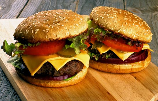 Homemade Hamburgers with Beef, Tomato, Lettuce, Pickle, Red Onion and Cheese into Sesame Buns closeup on Wooden Cutting Board