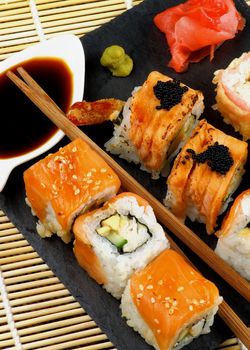 Arrangement of Various Maki Sushi with Smoked Salmon, Eel and Tempura Crab on Stone Plate with Ginger, Soy Sauce and Wasabi closeup on Straw Mat background