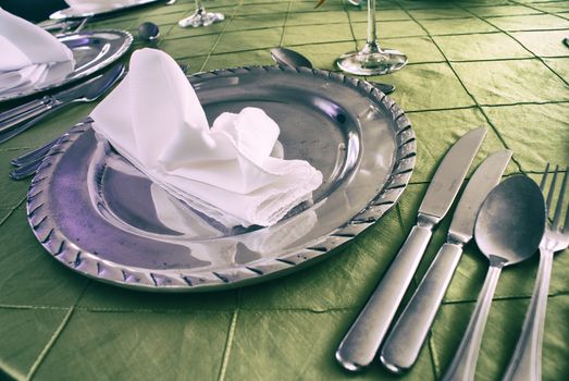 Photograph of some silver clutery and a plate with a napkin, on a green tablecloth