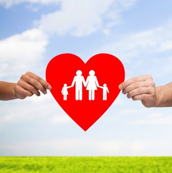 health, love, adoption and relationships concept - closeup of multiracial couple hands with big red heart with family and kids over blue sky with grass background