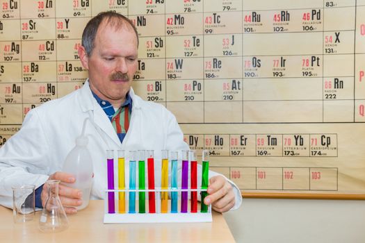 Chemist filling glass tubes with colored liquids in front of wallchart with periodic table