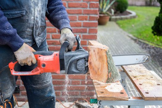 Man in work clothes holding chain saw cutting wood of trunk on table outdoors