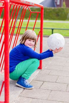 Young crouching dutch girl with white ball on hands in red metall goal on playground