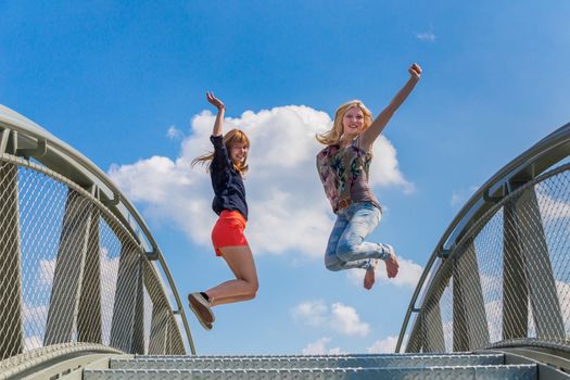 Two happy and enthusiastic teenage girls jumping on bridge in front of blue sky