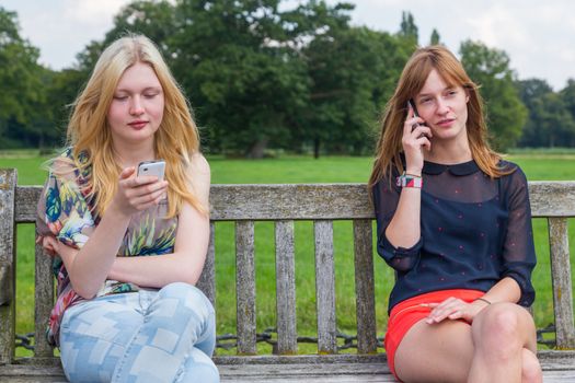 Two caucasian teenage girlfriends sitting on wooden bench in park calling with mobile phone