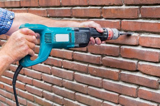 Arms of construction worker holding drilling machine against brick wall