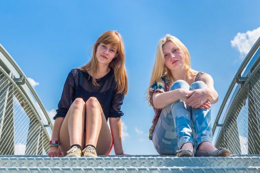Two dutch teenage girlfriends sitting on metal bridge with blue sky