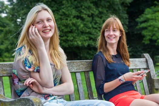Two european teenage girlfriends sitting on bench in park with mobile phone