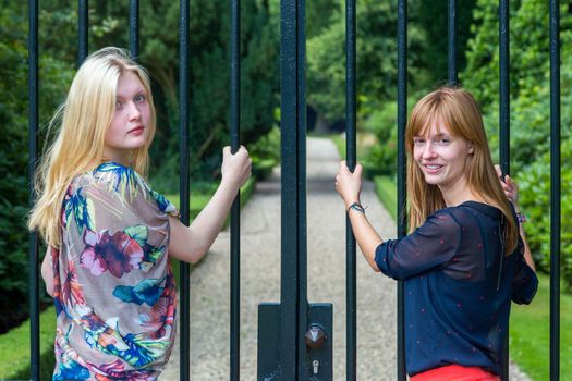 Two dutch teenage girlfriends holding metal bars of entry gate in park