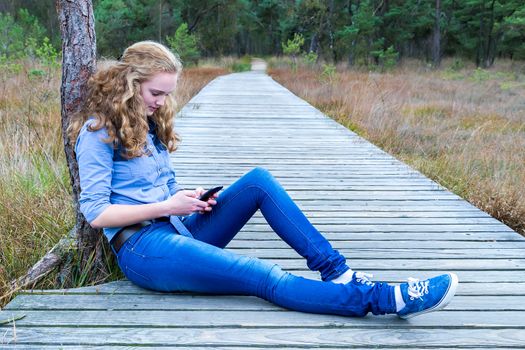 Caucasian teenage girl sitting on wooden footpath in nature phoning mobile