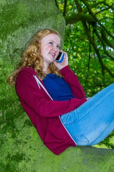 Dutch teenage girl calling with mobile telephone lying on branch of tree