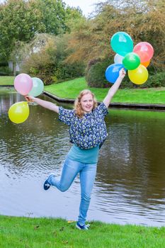European teenage girl holding up many  coloured balloons at water of pond