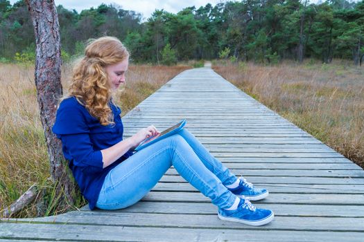 Caucasian teenage girl working with tablet computer on wooden footpath in nature